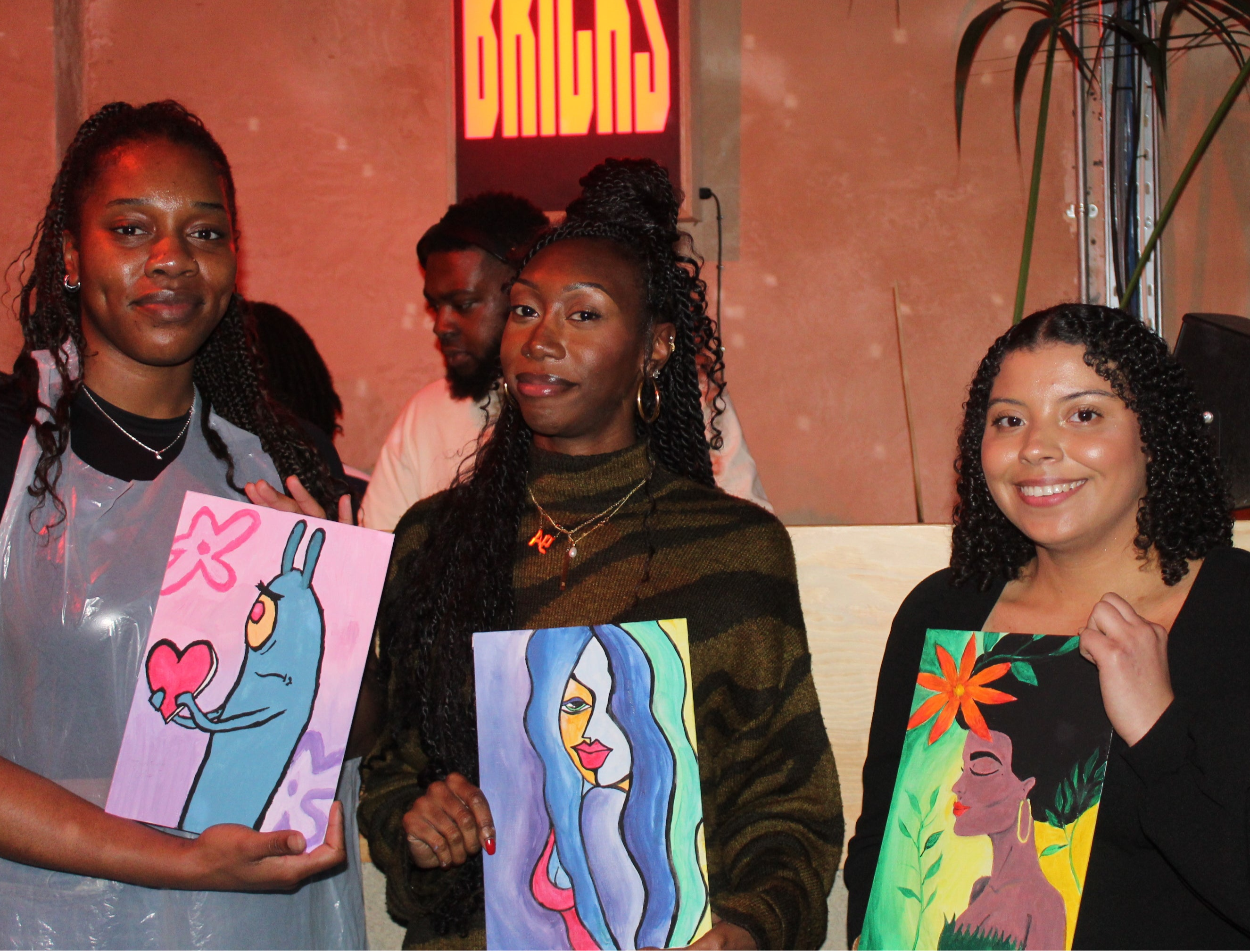 Three women holding colorful paintings indoors with a 'Briggs' sign in the background.